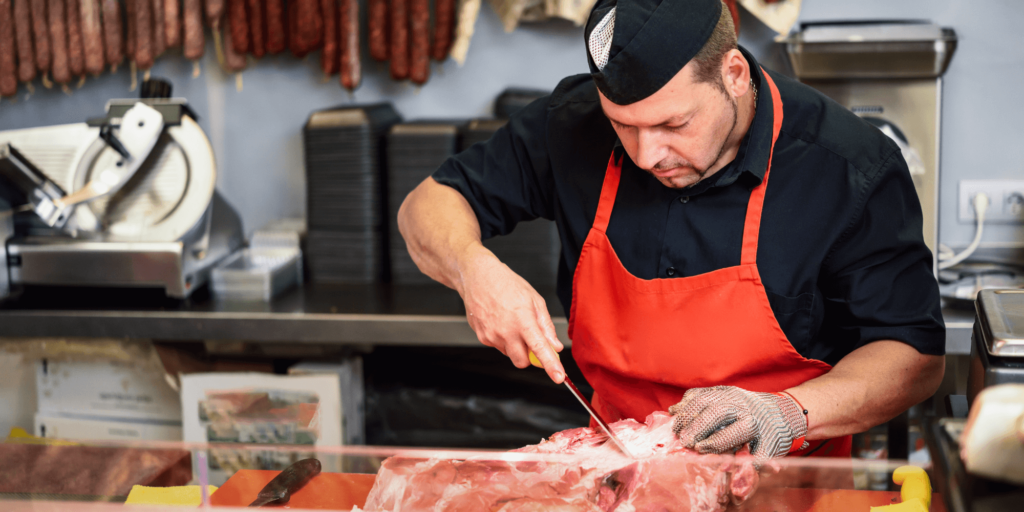 A professional butcher preparing fresh meat behind a display counter in a clean butcher shop.