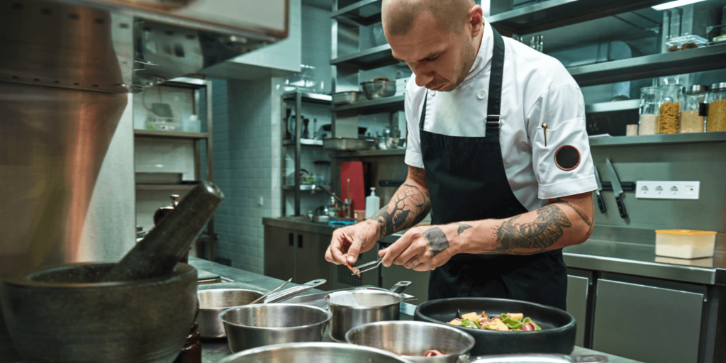 Focused chef in a professional kitchen preparing a meal with precision, surrounded by stainless steel equipment and cooking utensils, showcasing commercial catering operations.