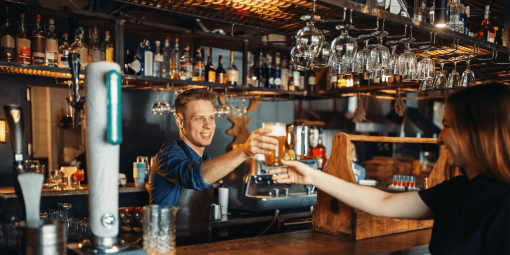 Bartender handing a pint of beer to a smiling female customer in a well-stocked pub bar with glasses and bottles in the background.