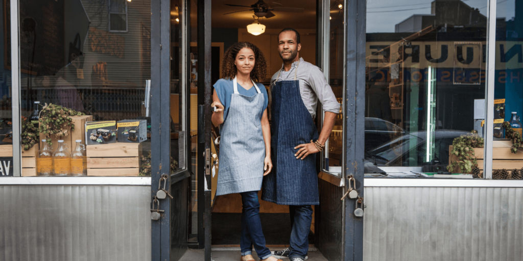 Two shop owners wearing aprons standing confidently in the doorway of their independent retail store, showcasing a welcoming and professional storefront.