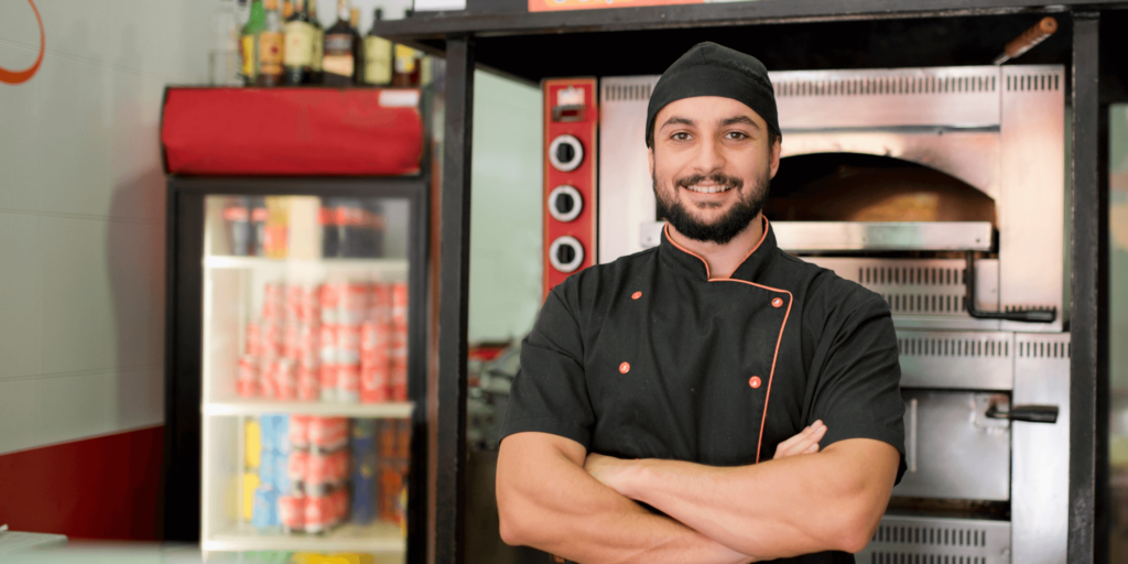 Smiling takeaway chef in uniform standing confidently with arms crossed in front of a commercial pizza oven and drinks fridge inside a fast-food shop.